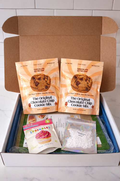 Open box containing cookie mix packages and additional baking supplies on a marble surface.
